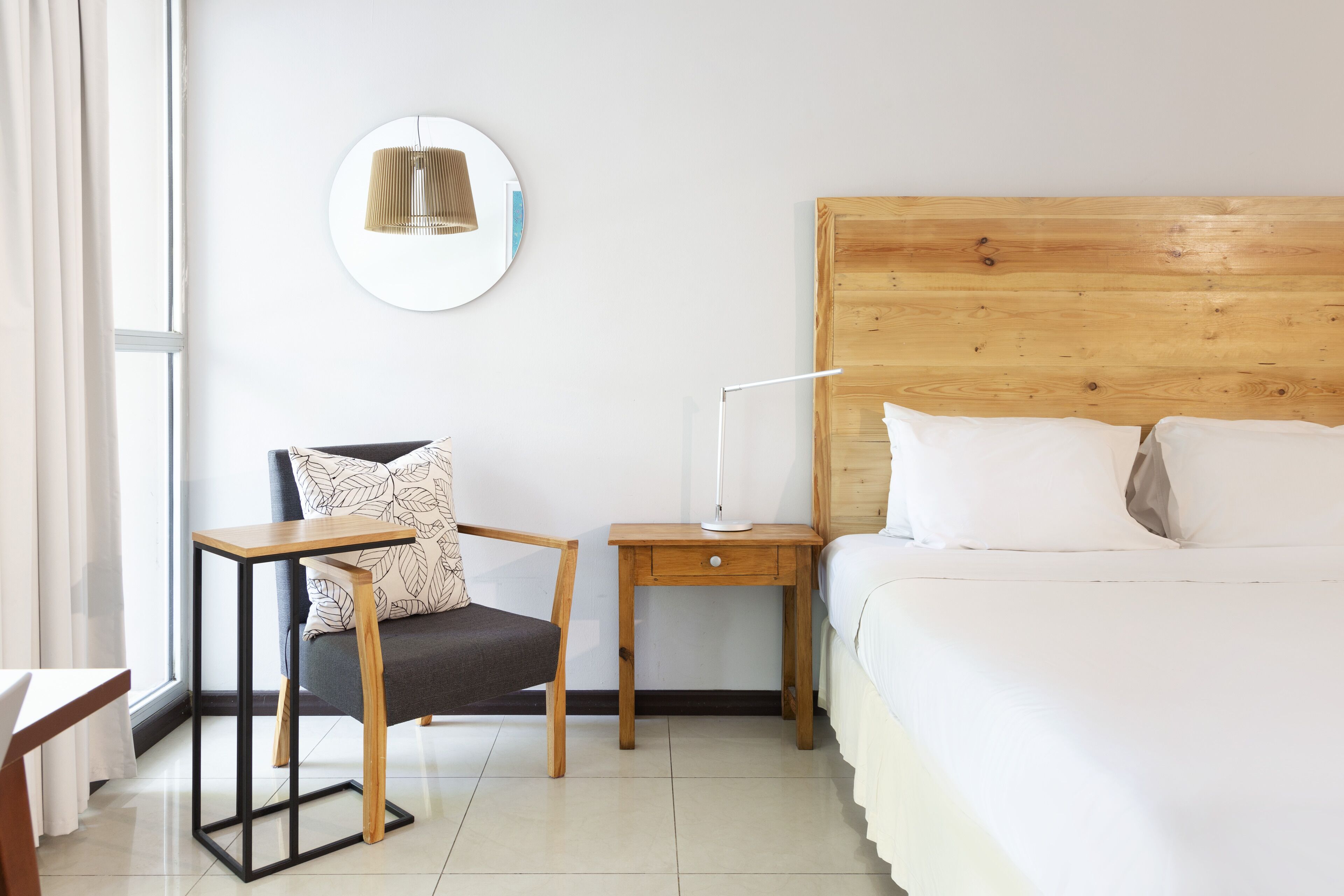 A hotel room with a wooden headboard, a bedside table with a lamp, a chair with a patterned cushion, and a large window.
