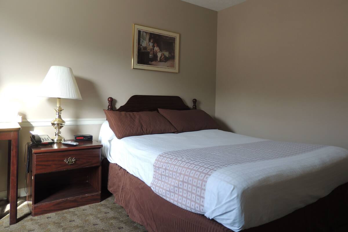 A neatly made bed with a patterned bedspread, a wooden nightstand with a lamp, a framed picture on the wall, and a small drawer unit.