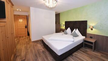 Habitación de hotel con suelo de madera, una cama grande con ropa blanca, cabecero de madera, mesita de noche, lámpara de pared y armario de madera.