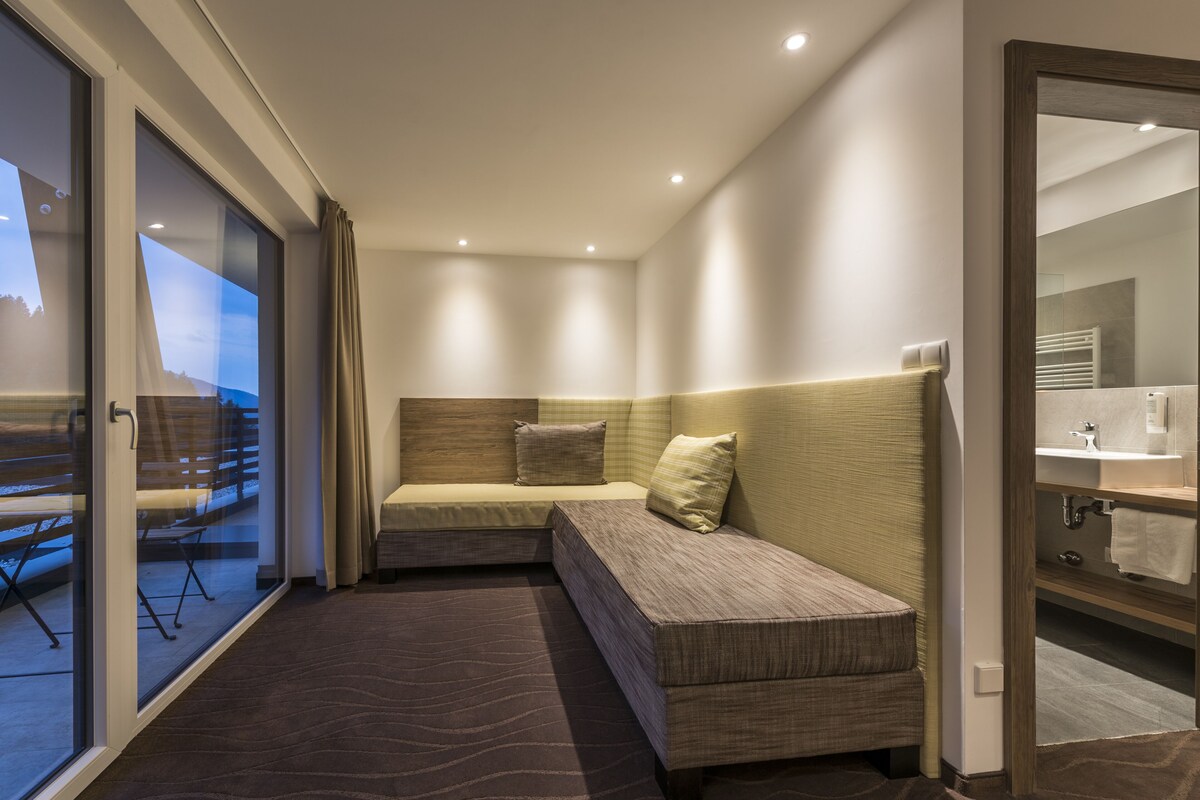 Una habitación de hotel moderna con un ventanal grande, un sofá cama, una mesita pequeña y un baño con espejo y lavamanos.