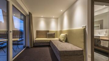 A modern hotel room with a large window, a sofa bed, a small table, and a bathroom with a mirror and sink.