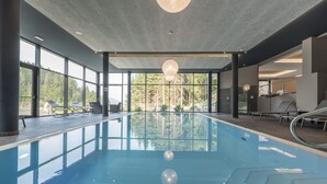 An indoor swimming pool with clear glass walls, modern lighting, and a view of a forested area. - Rechenmachers Rosengarten (Nova Levante)