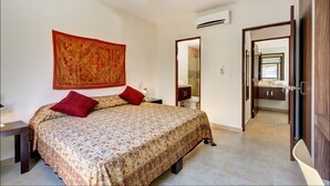 A bedroom with a large bed, two red pillows, a nightstand, a wall tapestry, an air conditioning unit, and a bathroom visible through an open door. - Ana's Village (Tulum)