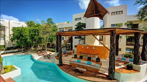 A swimming pool with a wooden deck and seating area, surrounded by buildings and trees. - Ana's Village (Tulum)
