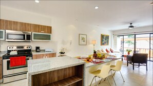 A modern kitchen with granite countertops, stainless steel appliances, and wooden cabinets. There is a dining area with a table and chairs, and a large window providing a view of the outdoors. - Ana's Village (Tulum)