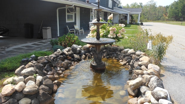 A stone fountain surrounded by rocks, with a house in the background.