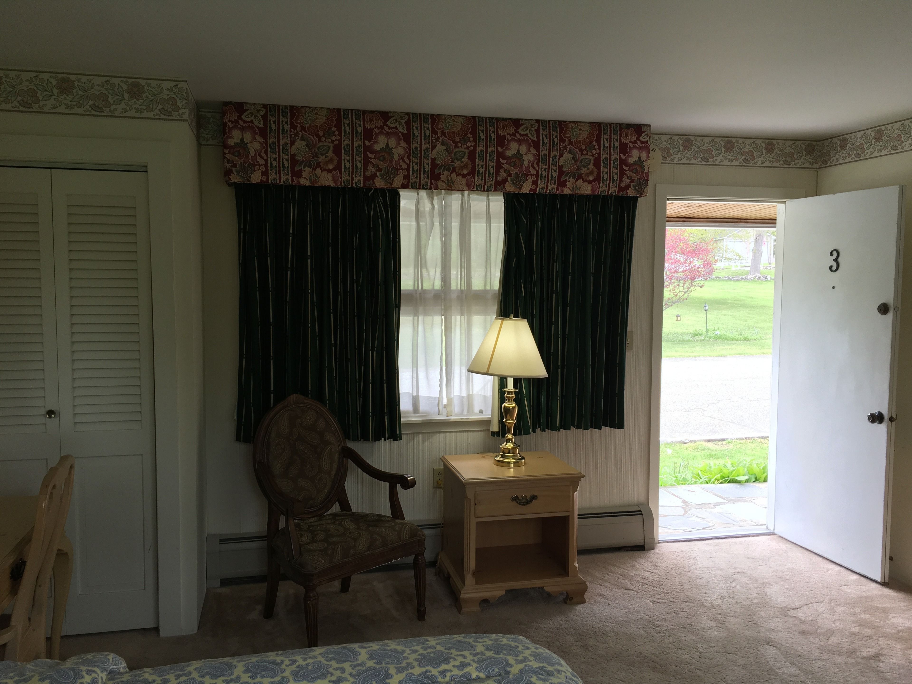 A room with a bed, a chair, a nightstand with a lamp, a window with dark curtains, and a door with the number 3.