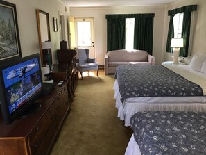 A hotel room with a television displaying a news channel, a sofa, two beds, a desk with a lamp, and a window with curtains.