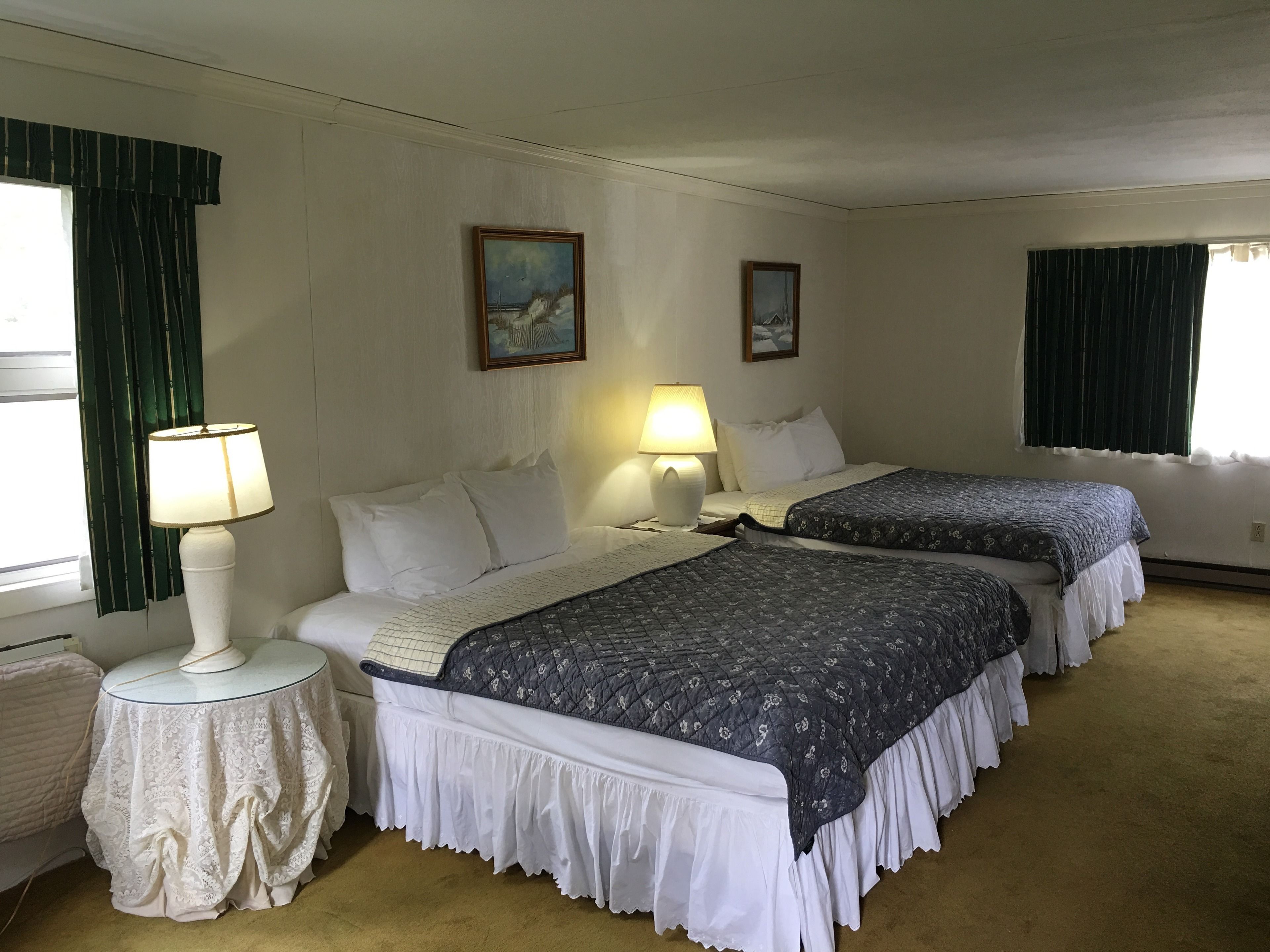A hotel room with two beds, a small table with a lamp, and two paintings on the wall.