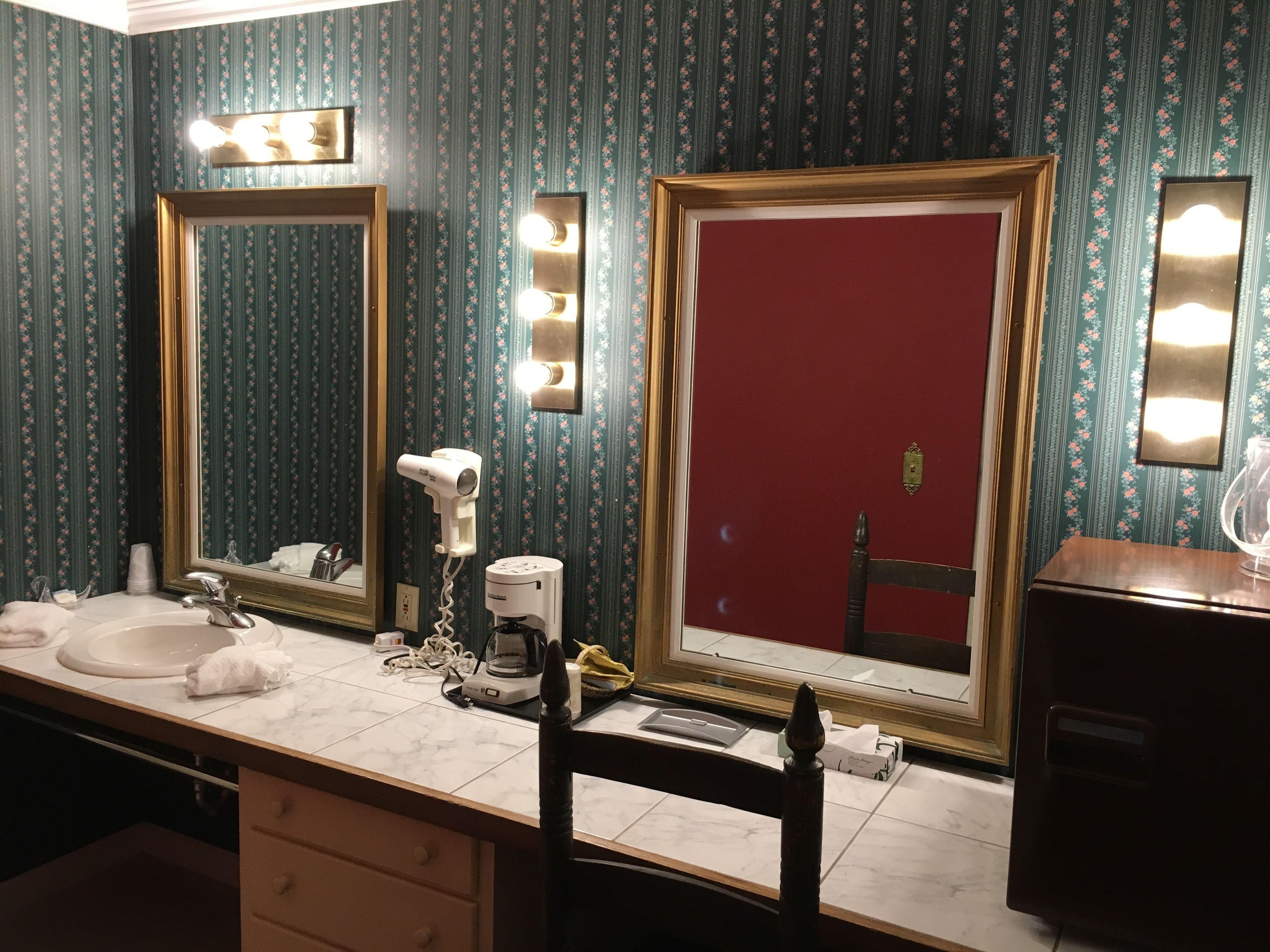A bathroom with two mirrors, a hairdryer, and a coffee maker on the counter.