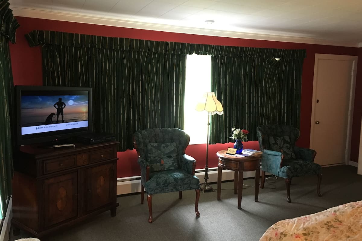 A hotel room with a television on a wooden cabinet, two patterned armchairs, a small round table with a vase, and a red wall.