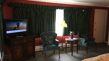 A hotel room with a television on a wooden cabinet, two patterned armchairs, a small round table with a vase, and a red wall.