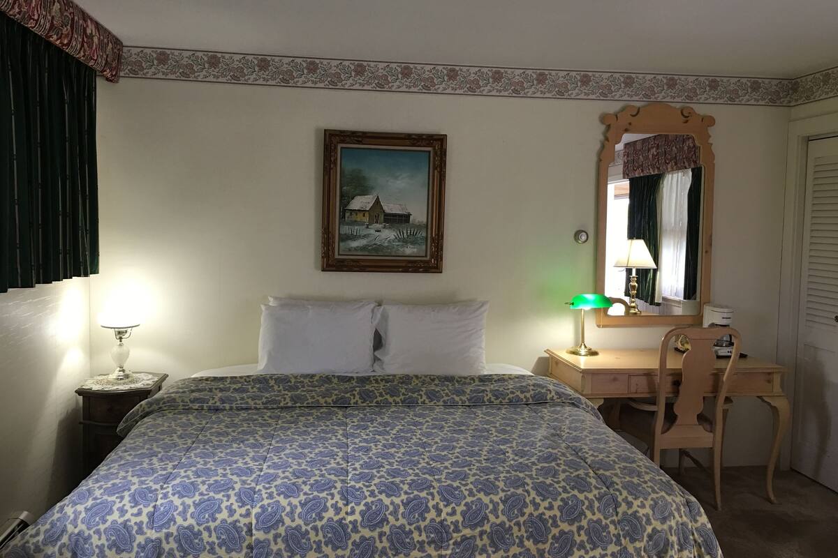 A hotel room with a bed, a desk, a chair, a lamp, a painting, and a mirror.