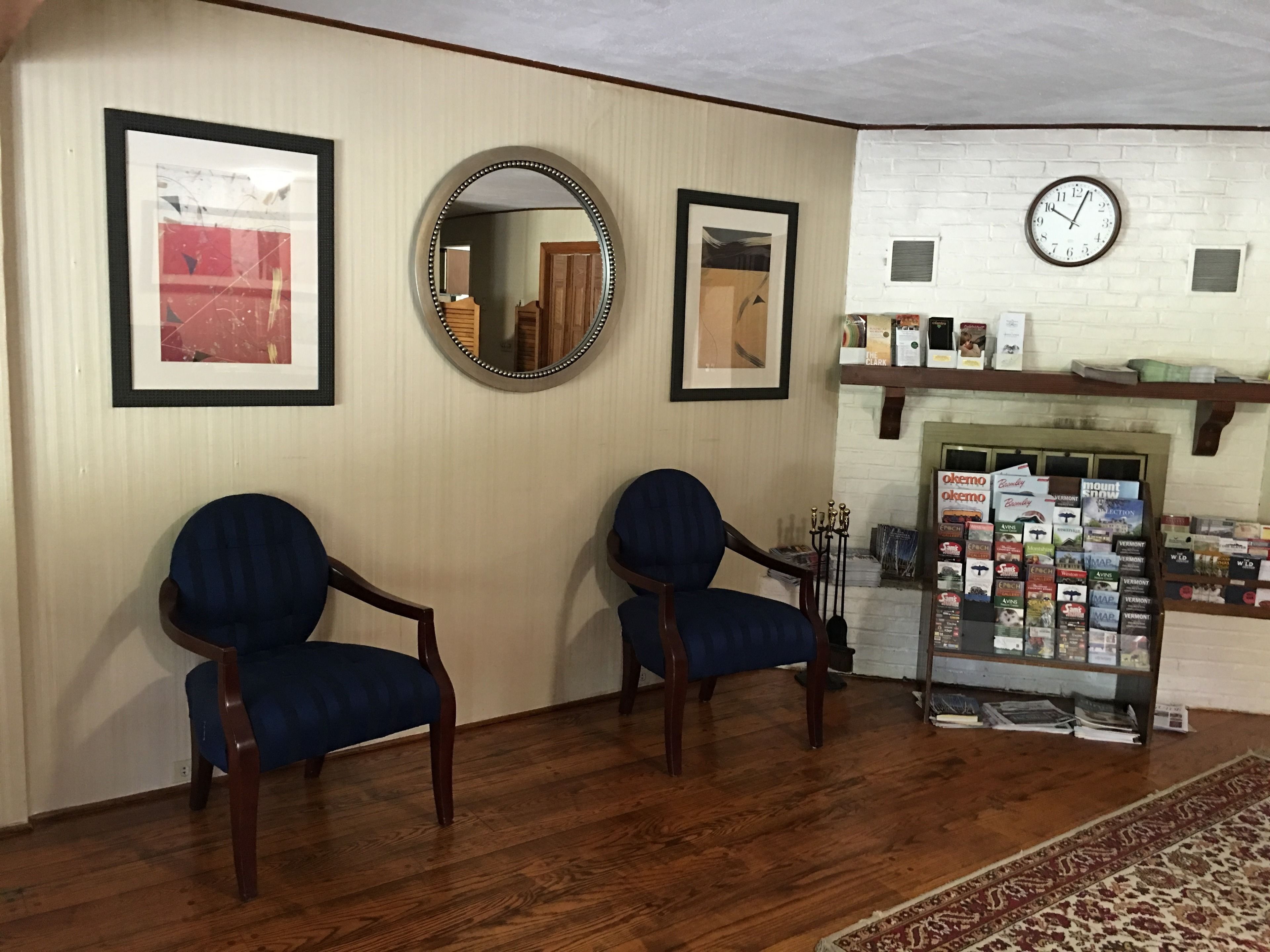 A reception area with two blue armchairs, a round mirror, framed pictures, a clock, and a fireplace with a bookcase.