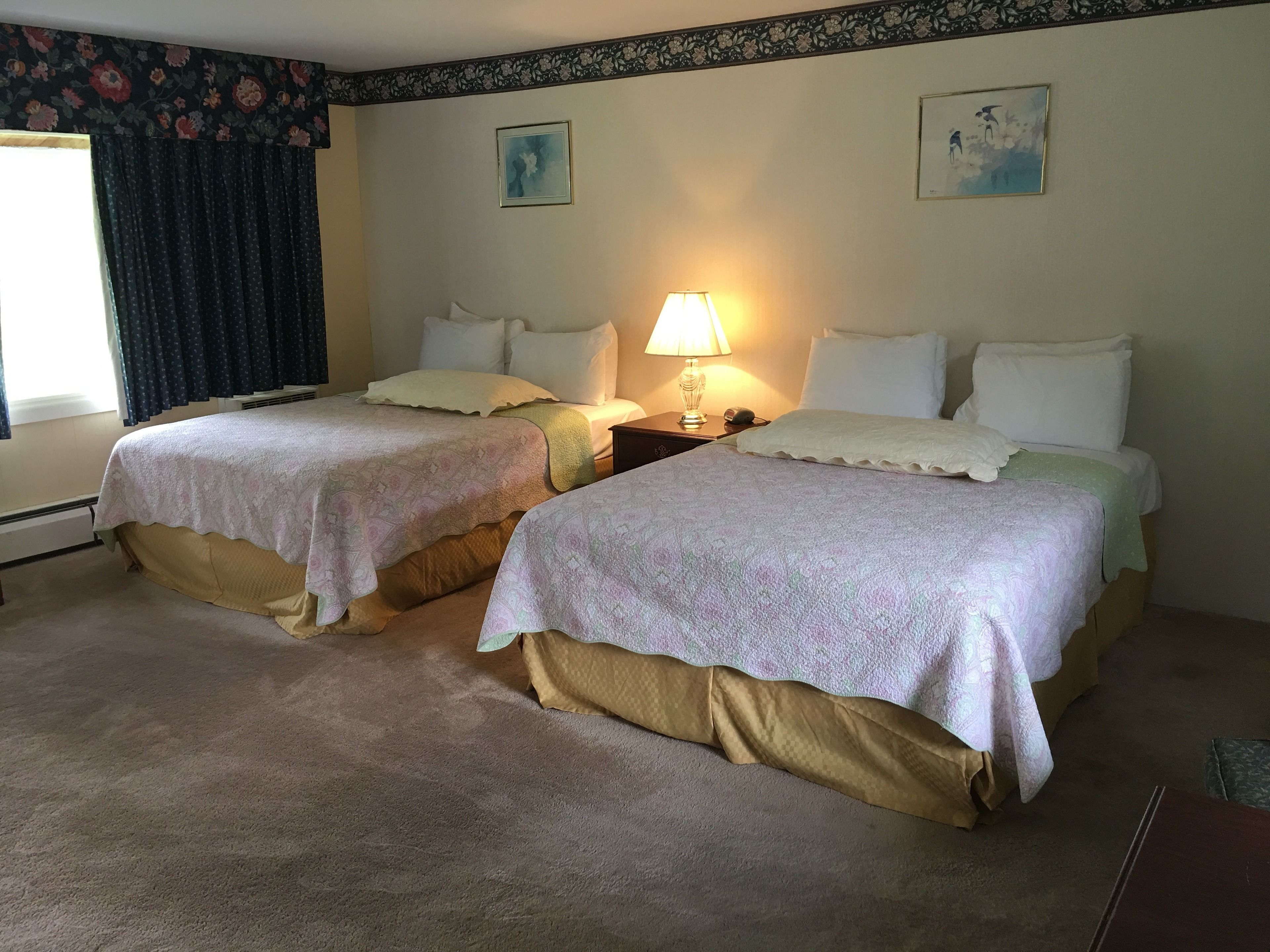 A hotel room with two beds, a nightstand with a lamp, floral wallpaper, and two framed pictures on the wall.