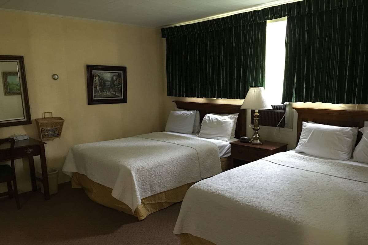 A hotel room with two beds, a nightstand with a lamp, a small table with a basket, and a framed picture on the wall.