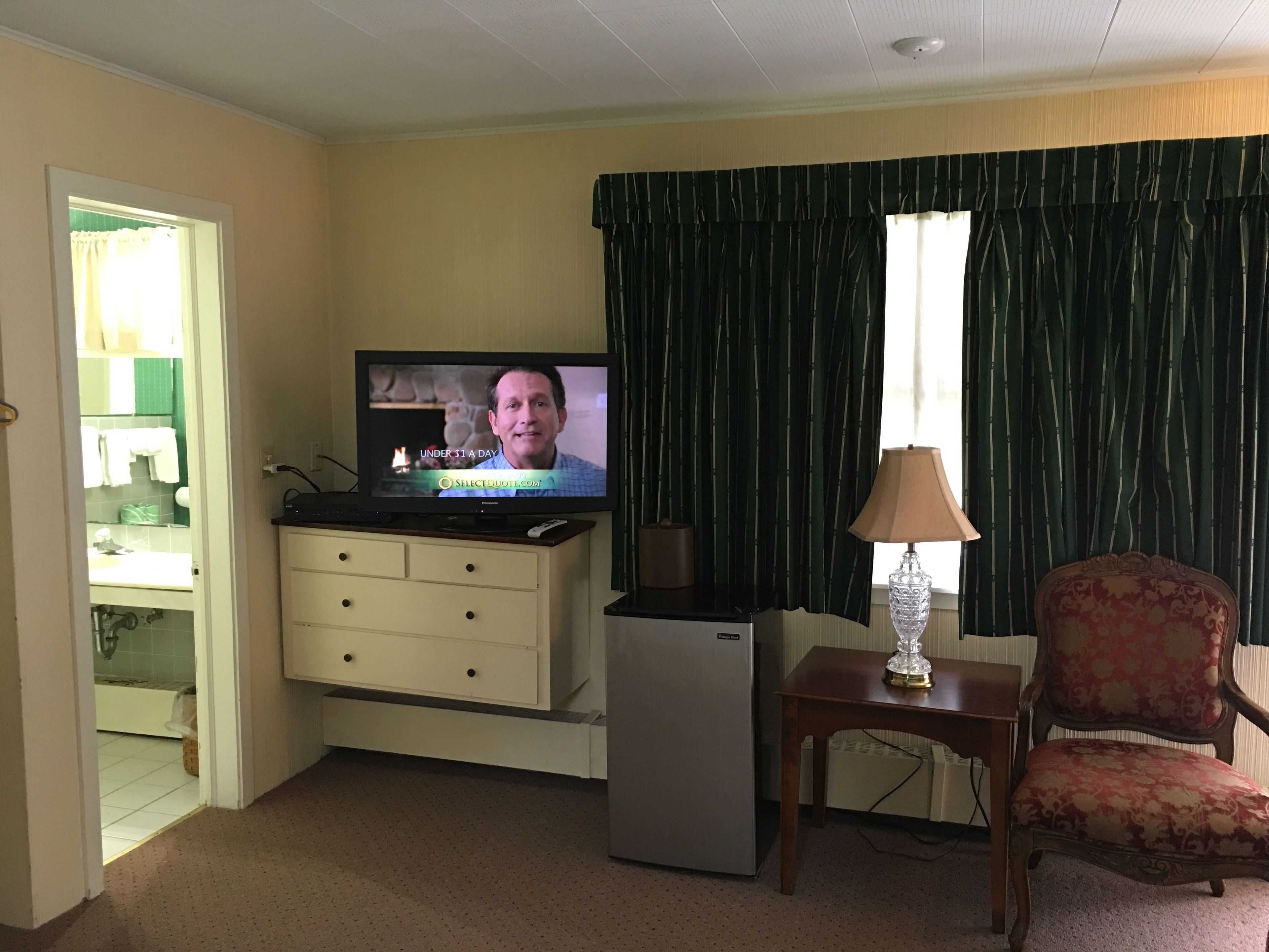 Una sala de estar con televisor, refrigerador, aparador de madera, una silla y una ventana con cortinas.