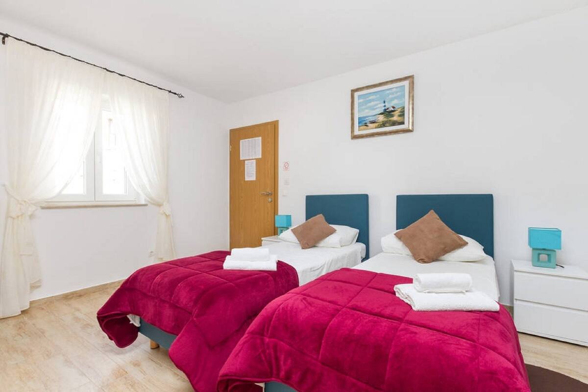 Hotellihuone, jossa on kaksi sänkyä. Jokaisessa sängyssä on punainen peitto ja valkoiset tyynyt. Huoneessa on puinen ovi, pieni yöpöytä, jossa on lamppu, sekä seinällä oleva kehystetty kuva.