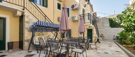 An outdoor patio with stone flooring, metal chairs, and tables, surrounded by a yellow building with balconies and air conditioning units.