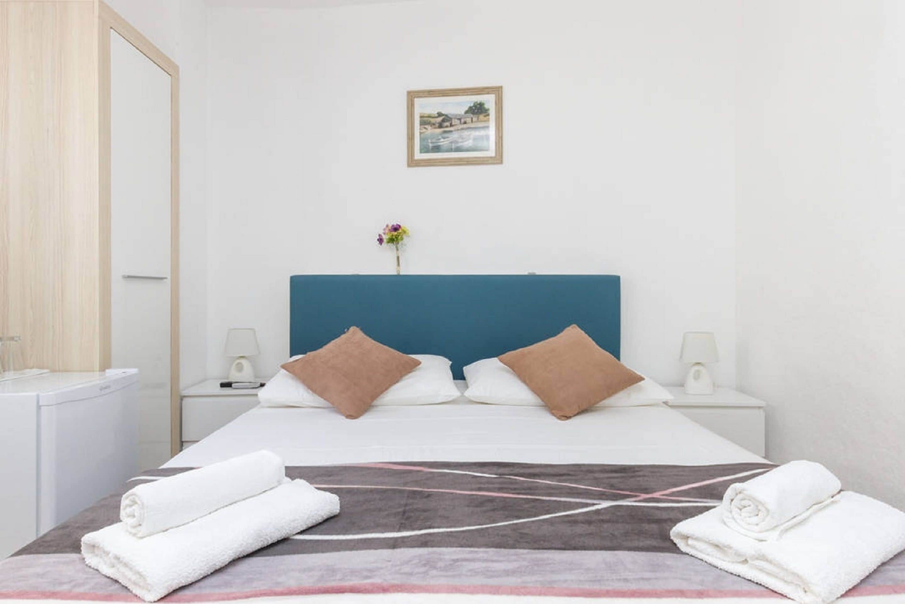 A hotel room with a bed, two pillows, a nightstand with a lamp, a framed picture on the wall, and a vase with flowers.