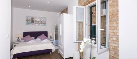 A modern bedroom with a bed, a white wardrobe, a window with a view, a small round table, and a painting on the wall.