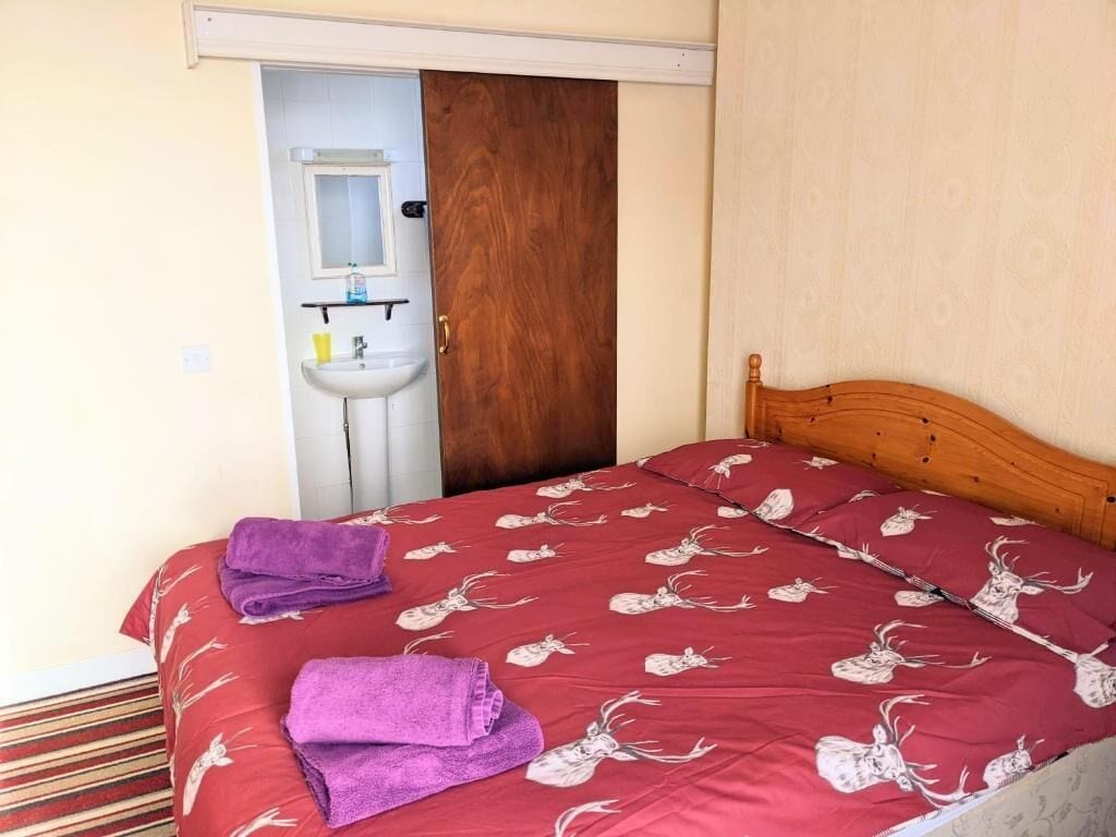 Ein Einzelbett mit einer rot-weißen Musterdecke, einem hölzernen Kopfteil und zwei gefalteten violetten Handtüchern auf dem Bett.