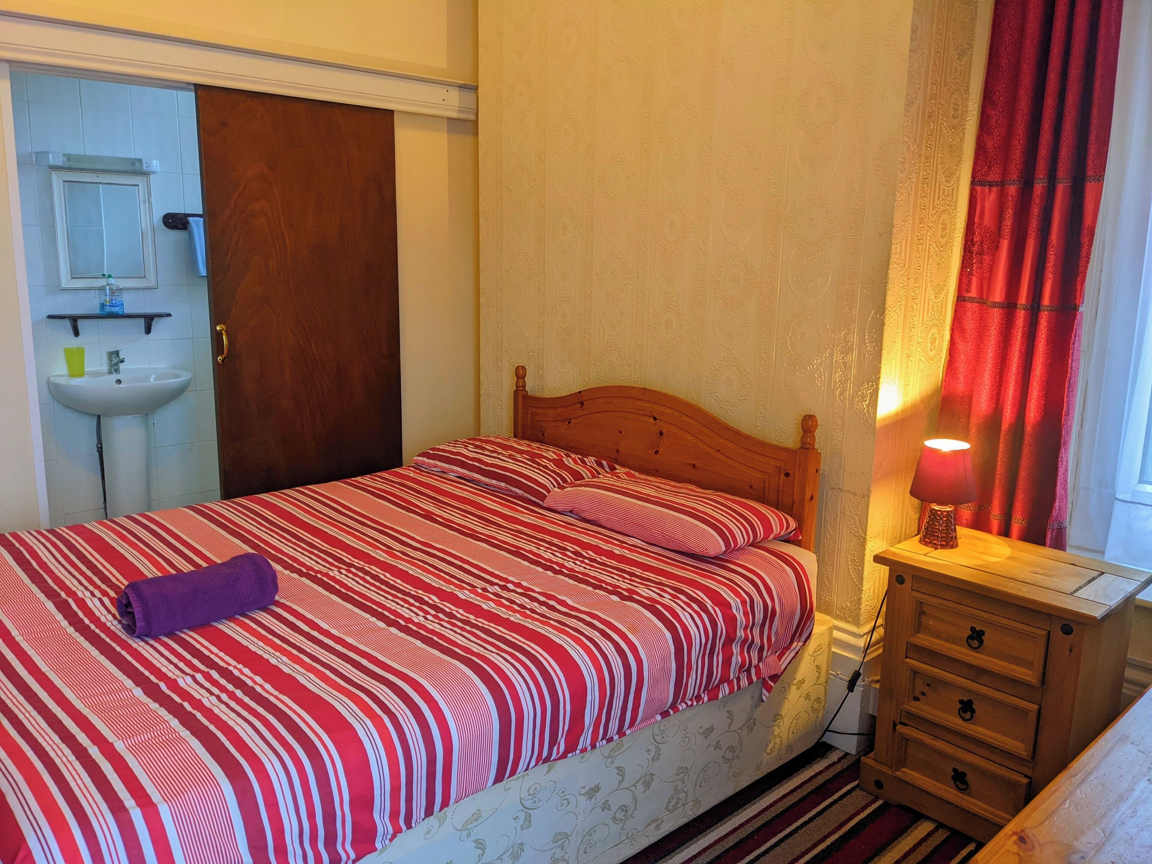 Een slaapkamer met een bed, een nachtkastje met lamp, een houten dressoir en een badkamer met wastafel en spiegel.