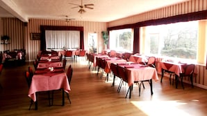 A dining area with tables set for multiple guests, a ceiling fan, and large windows with a view of trees. - The Altavia Hotel (Sandown)
