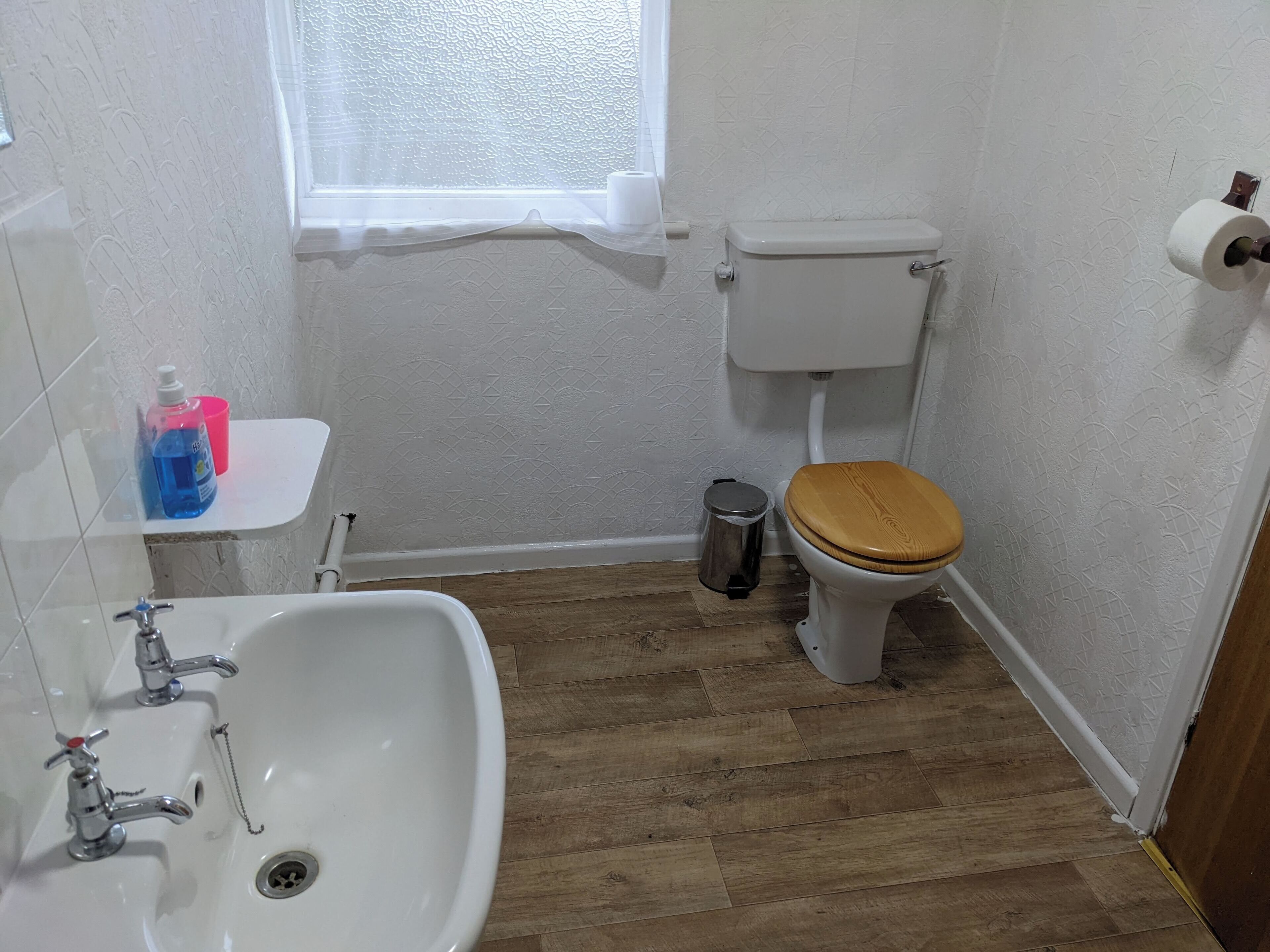 Et lille badeværelse med toilet i træ, hvid vask og et vindue.