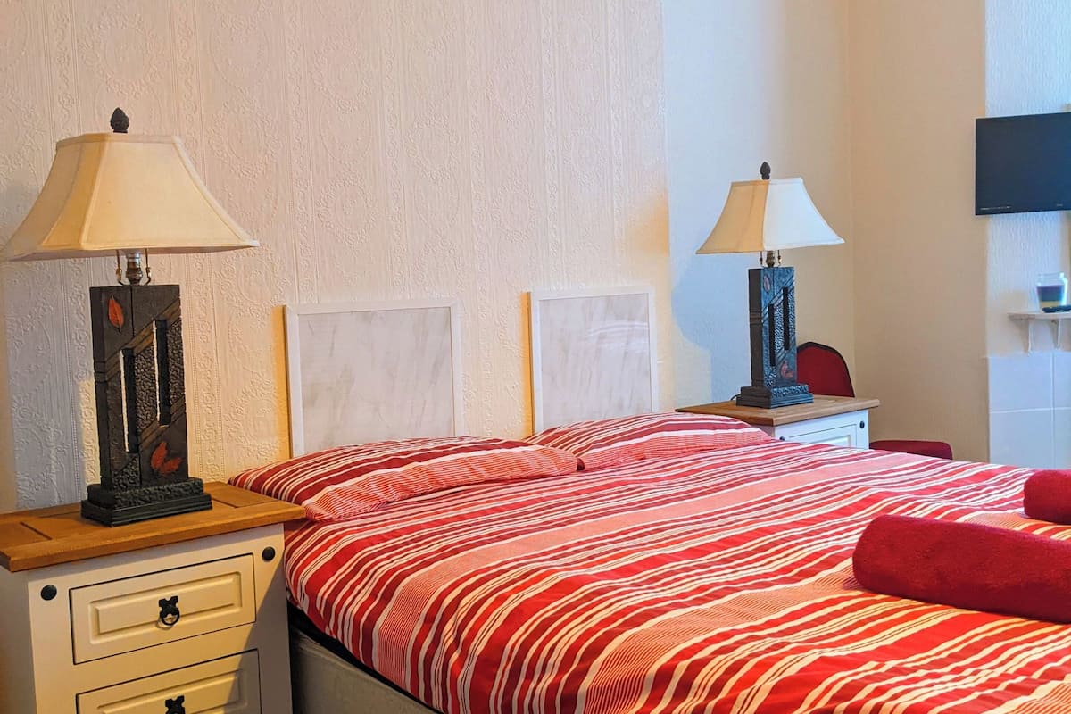 Uma cama bem arrumada com colcha listrada em vermelho e branco, duas mesas de cabeceira com abajures, um quadro na parede e uma televisão.