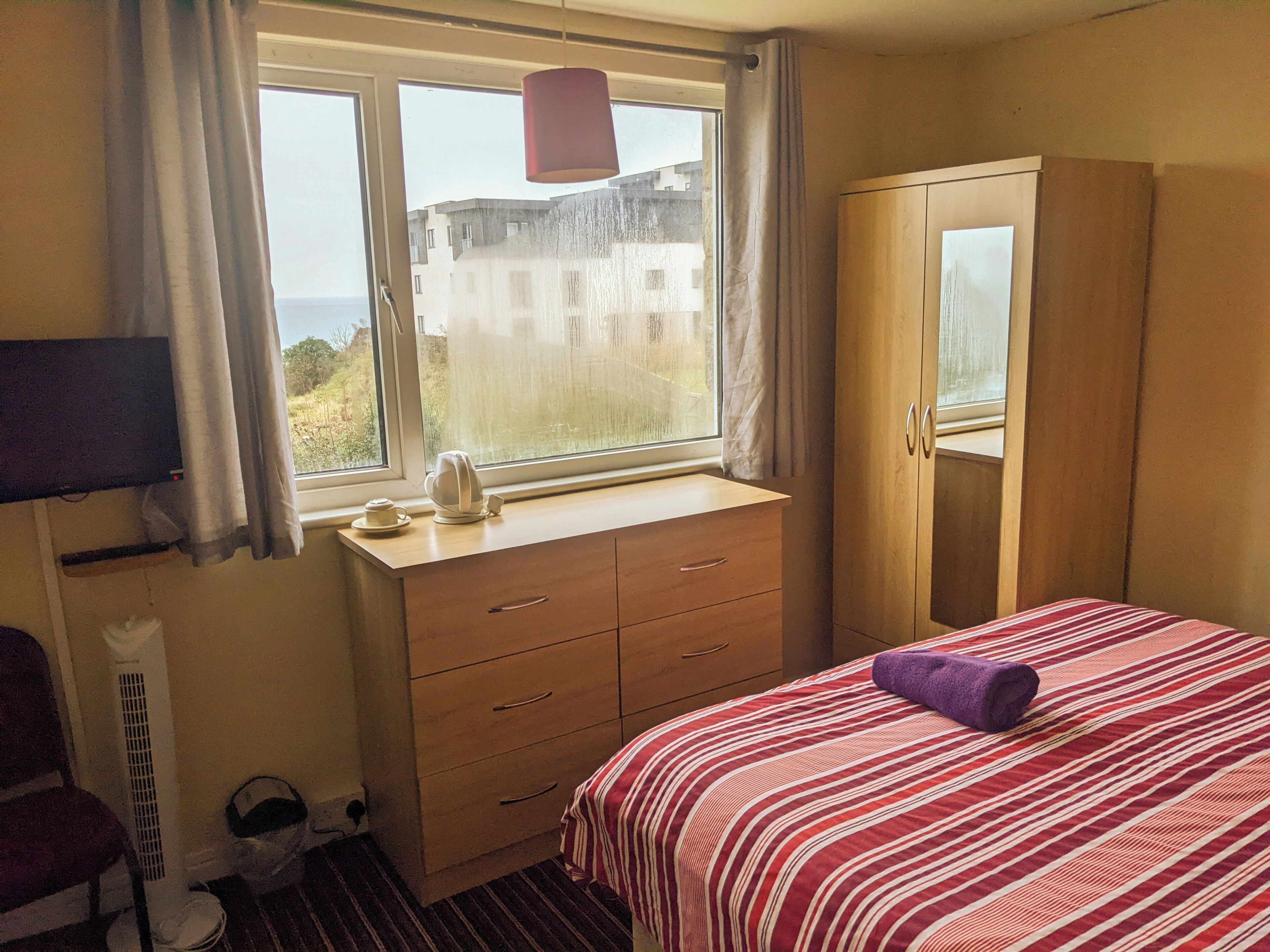 A bedroom with a bed, a wardrobe, a desk with a kettle, and a TV.