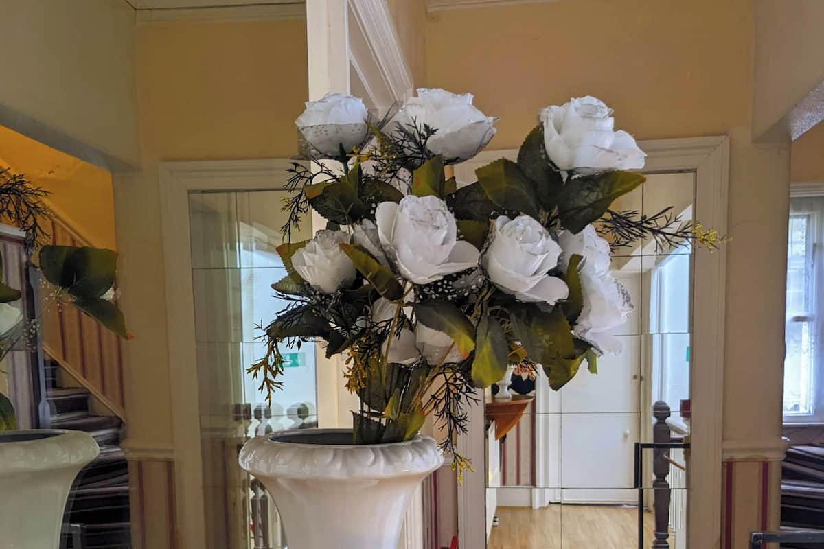 Um vaso branco com flores brancas sobre uma mesa de madeira em um quarto com escada e porta de vidro.