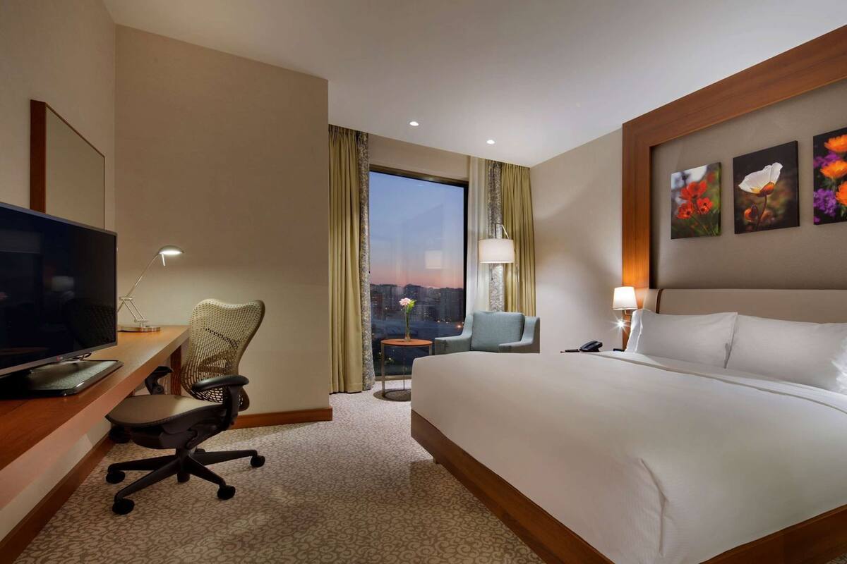 A hotel room with a bed, desk, chair, television, and artwork on the wall.