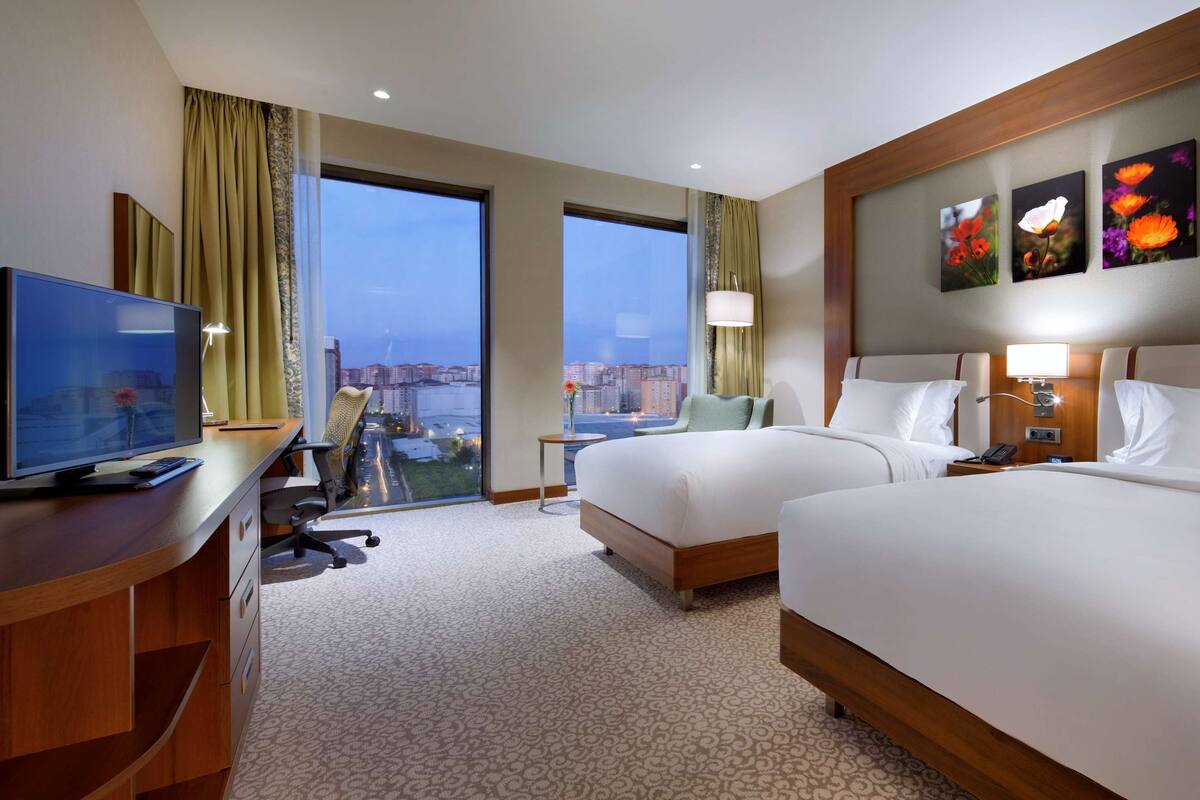 A hotel room with two beds, a desk with a TV, a desk lamp, a nightstand with a lamp, and a view of the city through large windows.