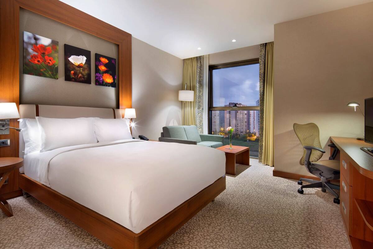 A hotel room with a large bed, a desk with a computer, a chair, and a window with a view of buildings.