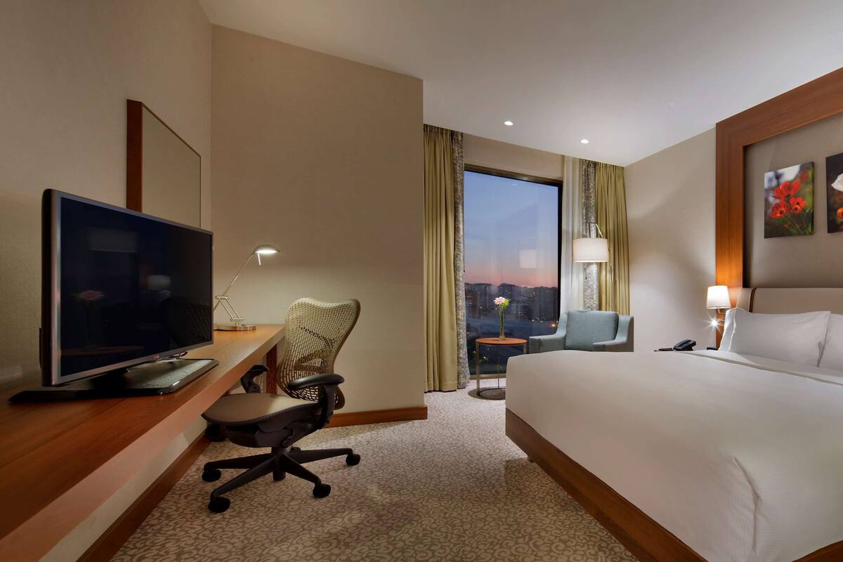 A hotel room with a large bed, a desk with a TV, a chair, a small table, and a view of the cityscape.