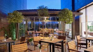 An outdoor dining area with wooden tables and chairs, surrounded by glass walls and plants. - Hilton Garden Inn Istanbul Beylikduzu (Istanbul)