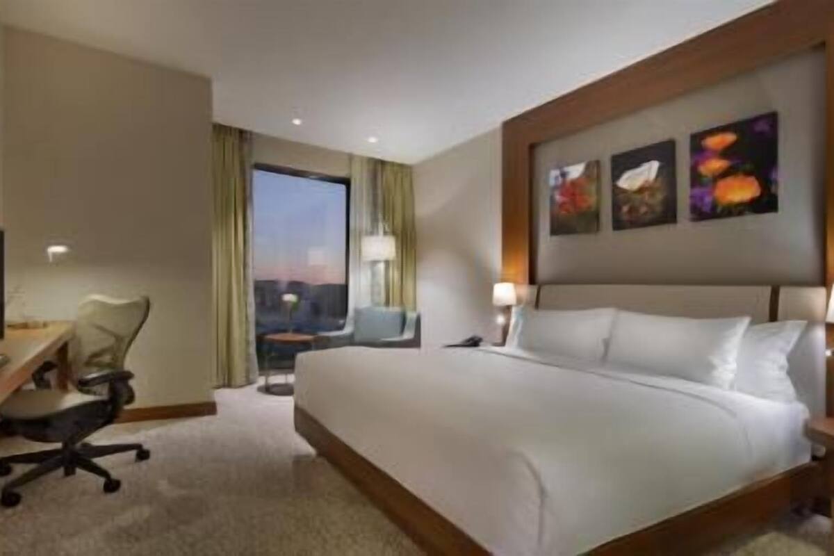 A hotel room with a bed, a desk with a TV, a chair, a nightstand, and a window with a view.