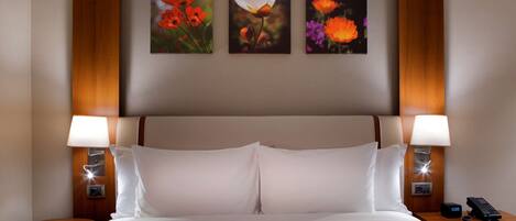A hotel room with a large bed, two bedside tables with lamps, and three framed floral paintings on the wall.
