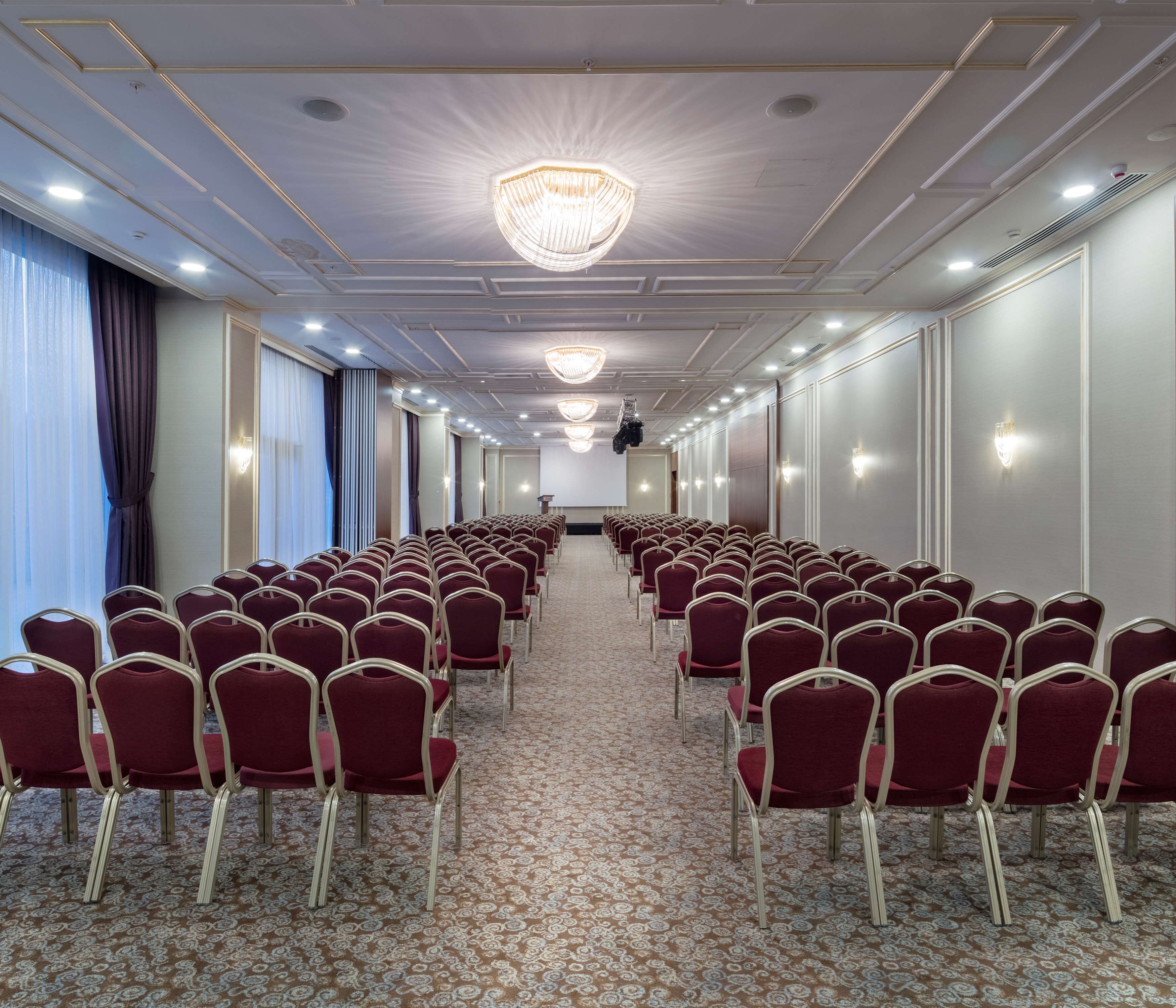 Ein Konferenzraum mit Reihen roter Stühle, einem gemusterten Teppich und einer Kronleuchter.