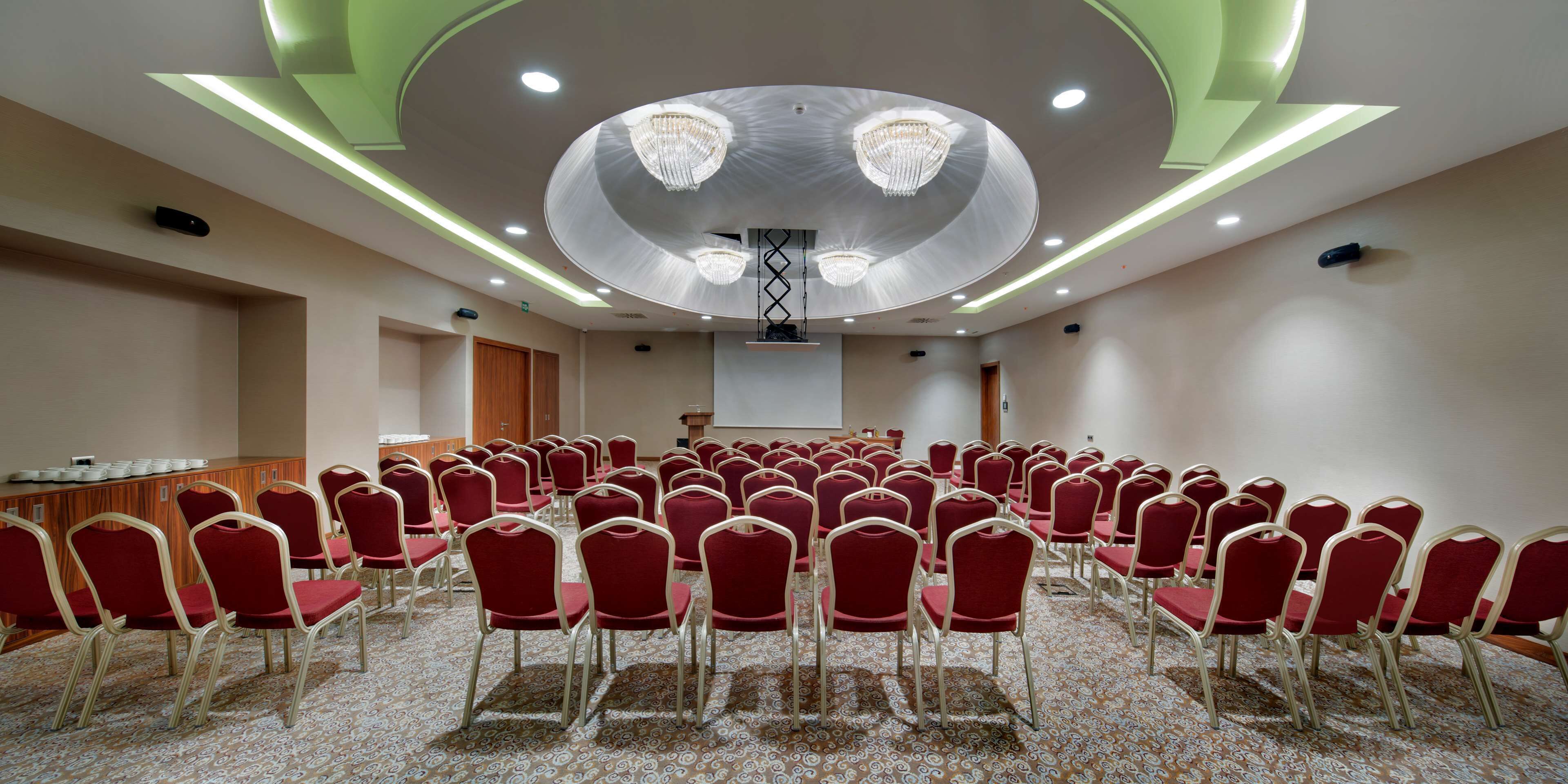 A conference room with rows of red chairs, a podium, and a large circular ceiling design with chandeliers.