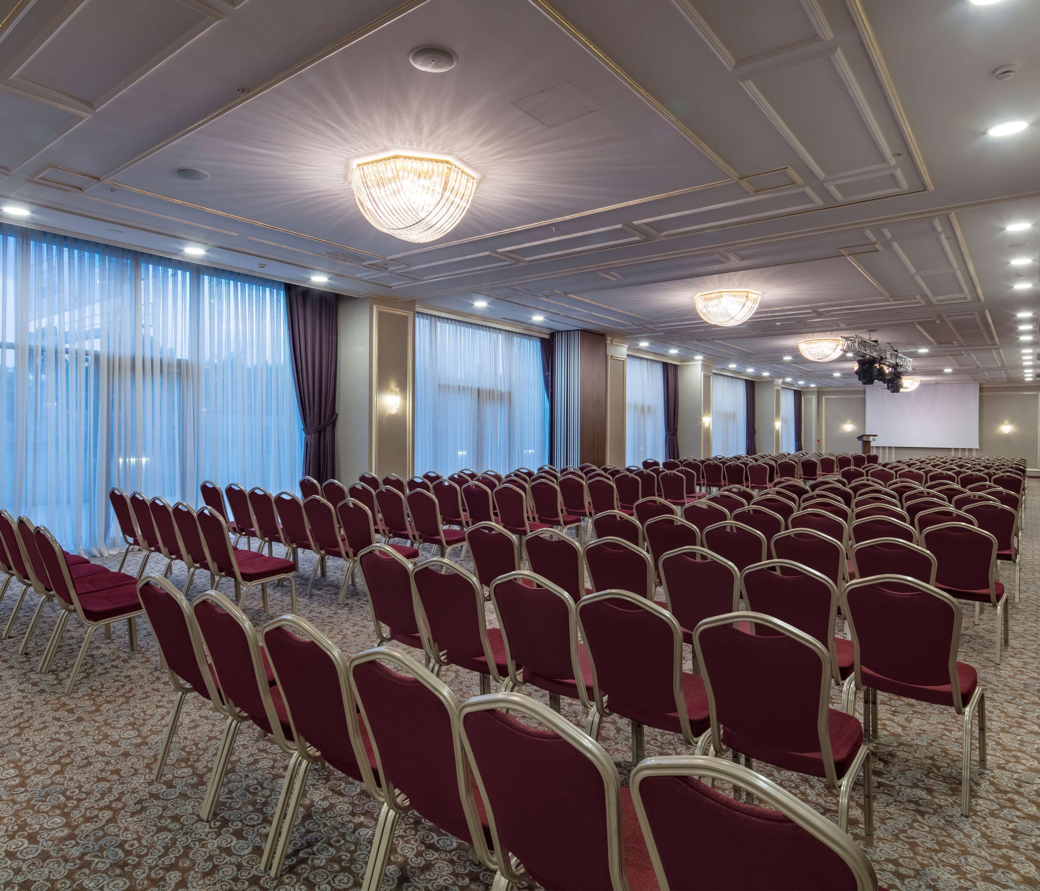 A conference room with rows of red chairs, large windows with sheer curtains, and a patterned carpet.