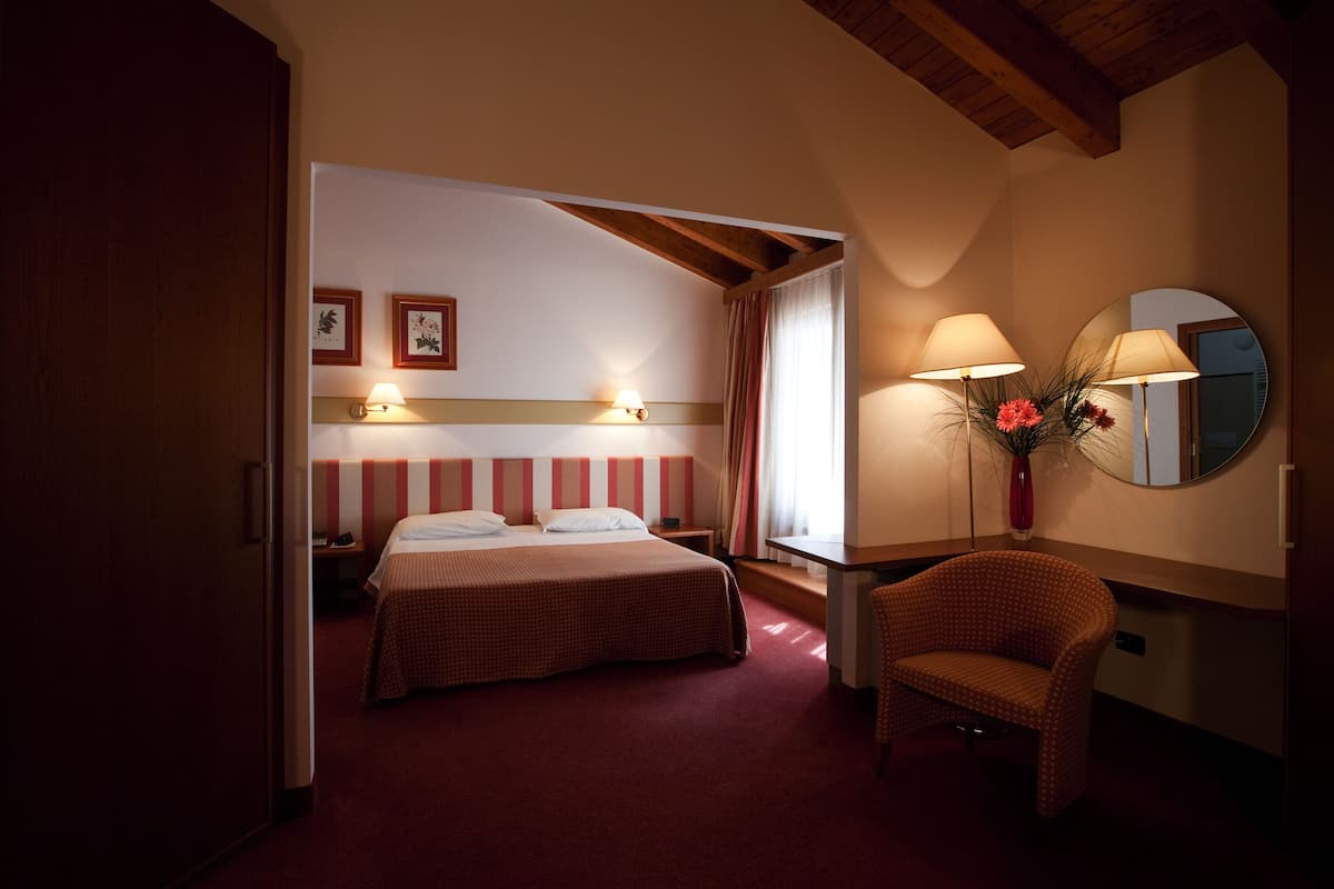 Habitación de hotel con cama, una silla, escritorio, espejo y dos lámparas fijadas a la pared.