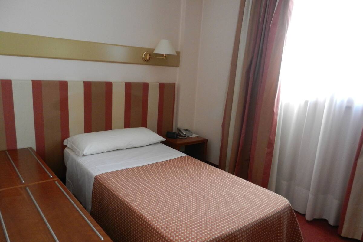 Una habitación de hotel con una cama, una mesita de noche de madera, una lámpara y una ventana con cortinas.