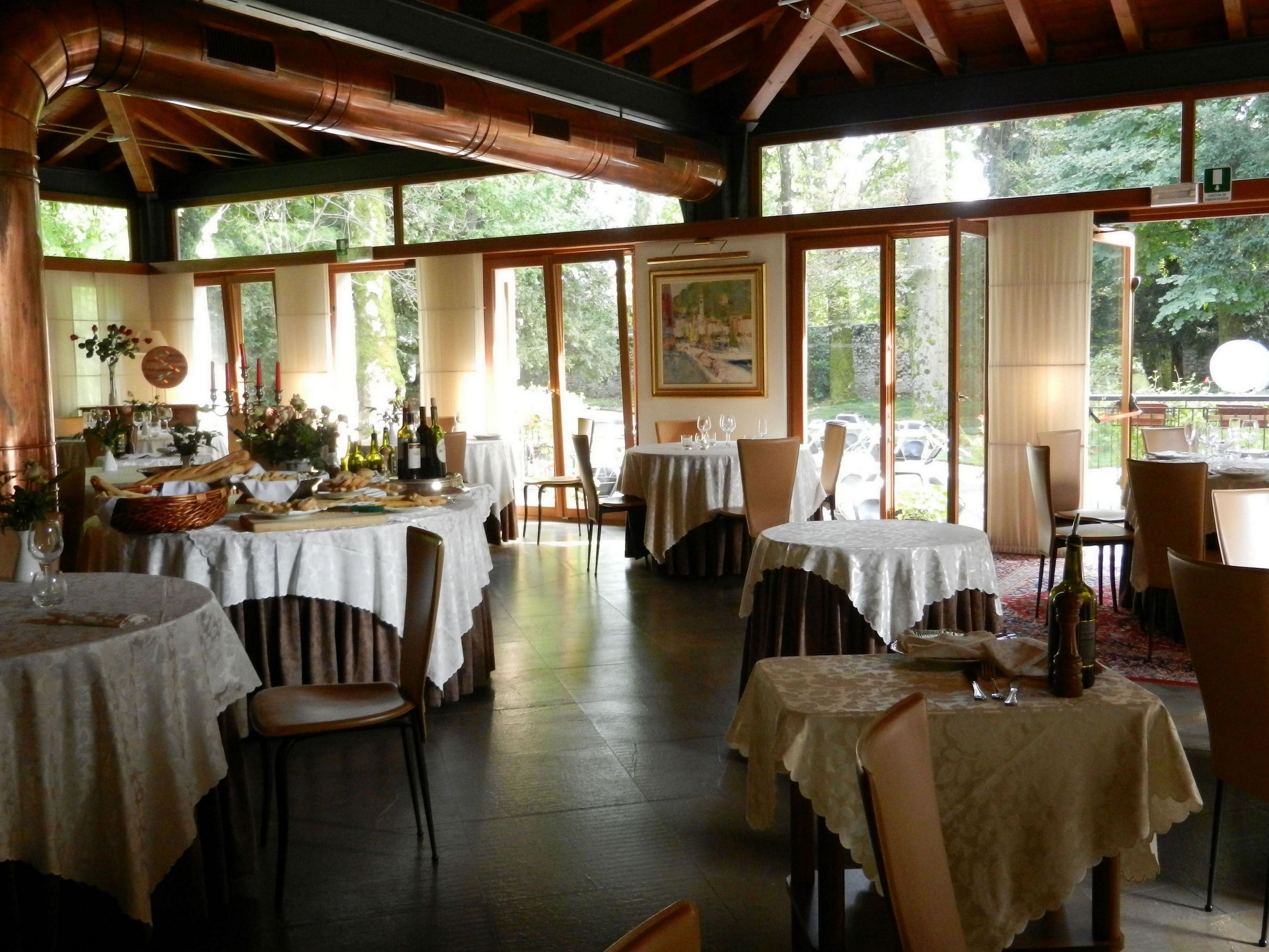 Ein Restaurant mit mehreren gedeckten Tischen, großen Fenstern mit Blick ins Freie und einer rustikalen Holzbalkendecke.