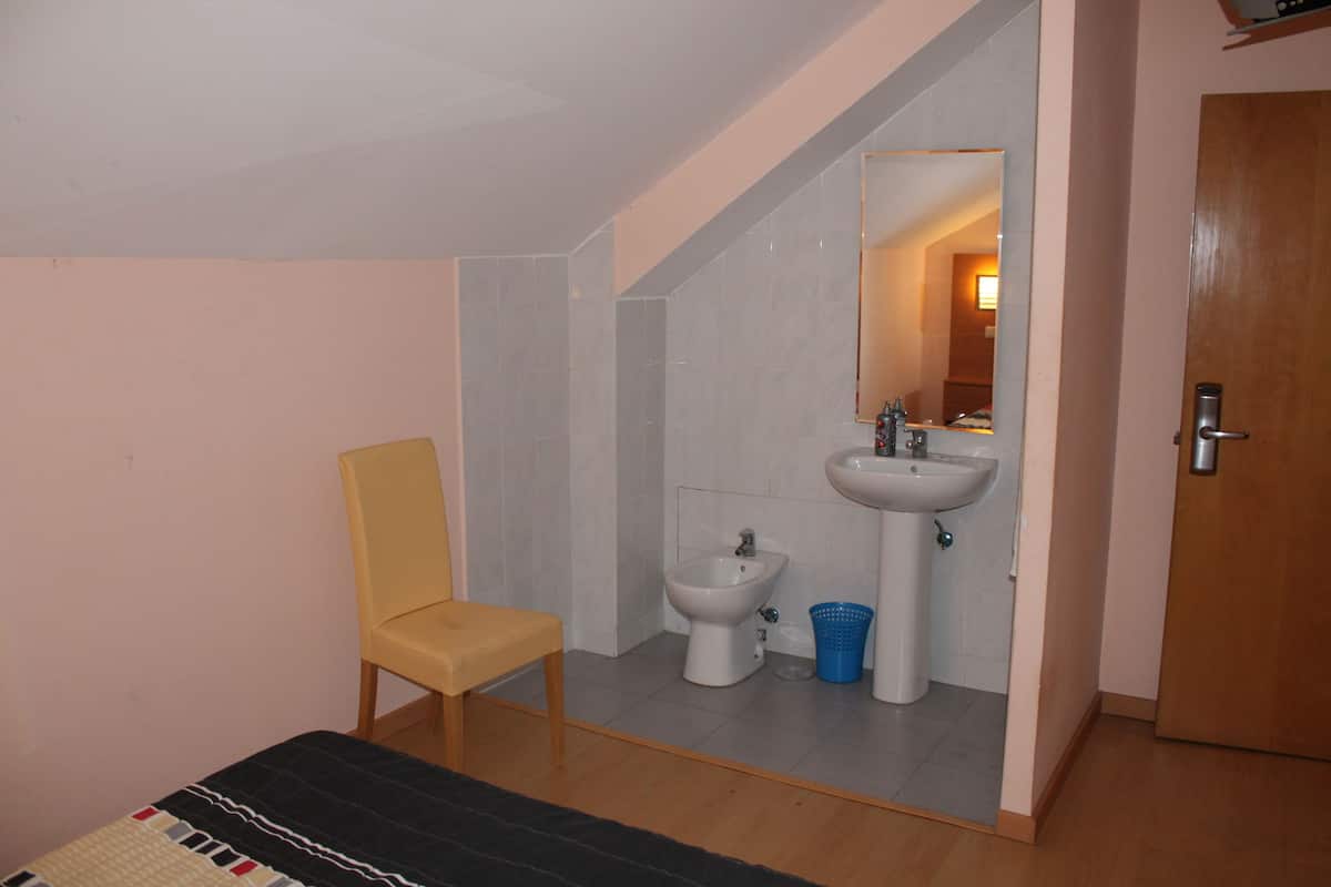 A small bathroom with a toilet, bidet, sink, and mirror. There is a chair and a bed in the room.