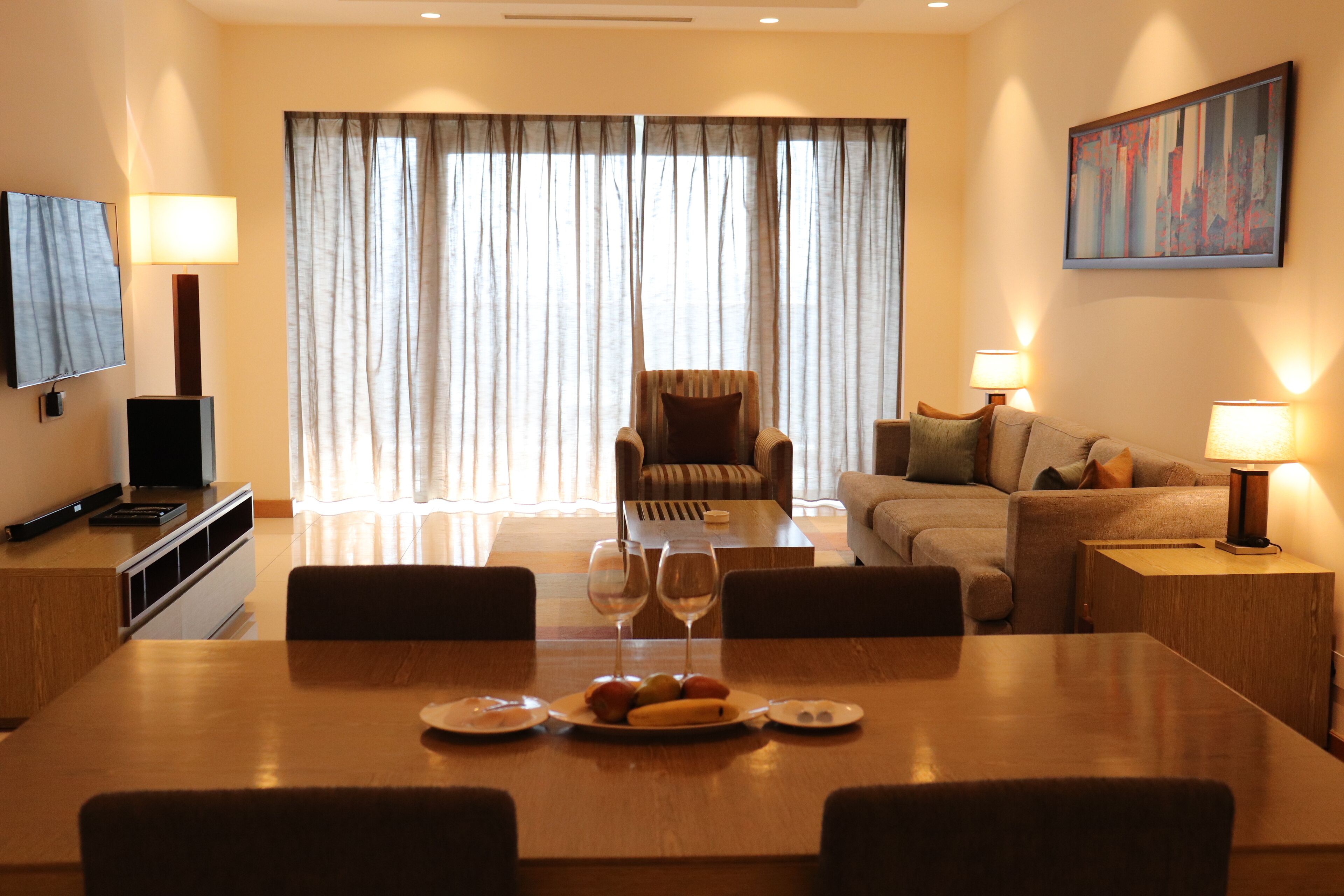 A dining area with a wooden table set for two, two wine glasses, and plates with food. There is a television on a stand to the left, a sofa and armchairs in the background, and a painting on the wall.