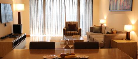 A dining area with a wooden table set for two, two wine glasses, and plates with food. There is a television on a stand to the left, a sofa and armchairs in the background, and a painting on the wall.