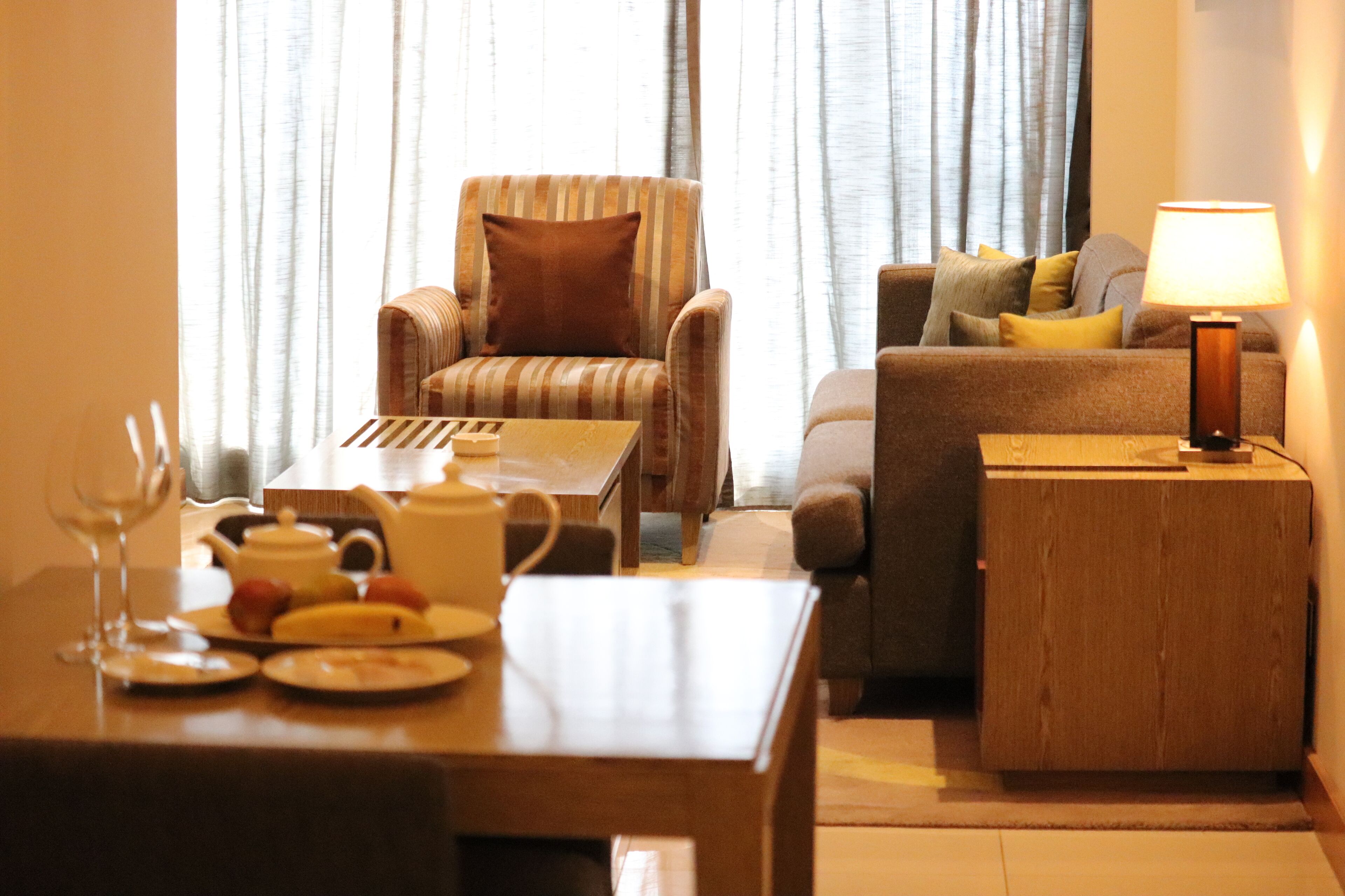 Una sala de estar con un sillón a rayas, un sofá con cojines, una mesita de madera con una lámpara y una mesa de comedor con platos y una tetera.