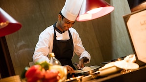 A chef in a white uniform and hat, preparing food at a counter with a red lamp overhead. - Oakwood Residence Kapil Hyderabad (Hyderabad)